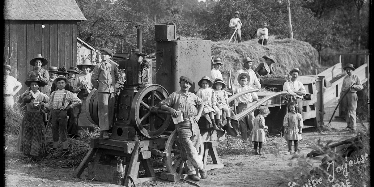 Photographie de l’équipe de Jules Boisramé lors du battage du blé chez Louis Roussel, entre 1920 et 1922, au Val-d’Izé. Une équipe d’environ vingt personnes, mêlant femmes, hommes et enfants. Jules Boisramé est accoudé à la batteuse (au centre). Photographe : Jean Hervagault (1879-1954)